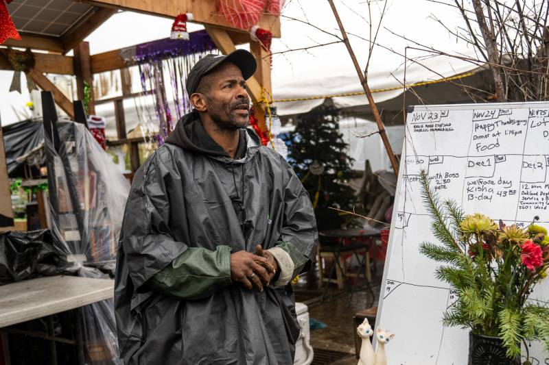 A man wearing a hat and rain jacket holds his hands together next to a whiteboard calendar with Christmas tree in the background.