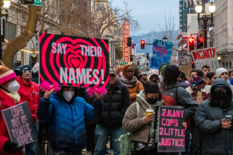 A sign that reads "Say their names!" is held by a person in a crowd of people holding signs.