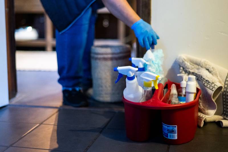 a person is seen from the waist down wearing blue rubber gloves picking up cleaning supplies from a red bucket