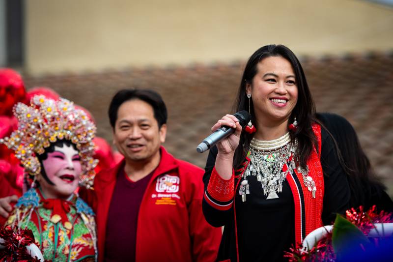 A woman wearing red and black clothing holds a microphone outdoors next to other people dressed in red.