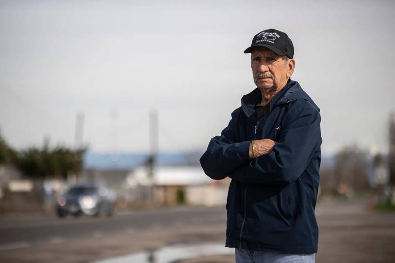 An older Latino man with a blue shirt and a blue cap looks at the camera with arms crossed, looking worried, houses blurred out in the background.