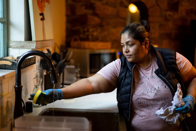 a Latina woman in a pink shirt and rubber gloves cleans a kitchen counter with a sponge