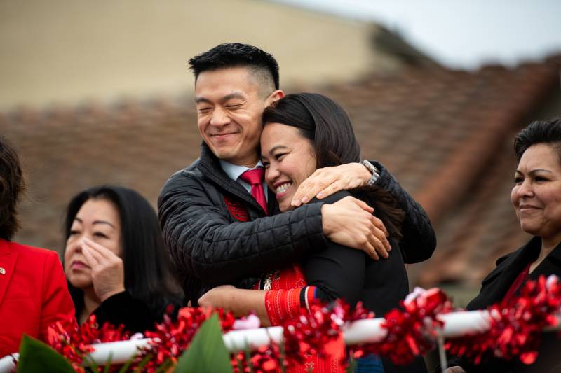 A man and woman, both Asian American, smiling happily, and wearing black clothing, embrace outside.