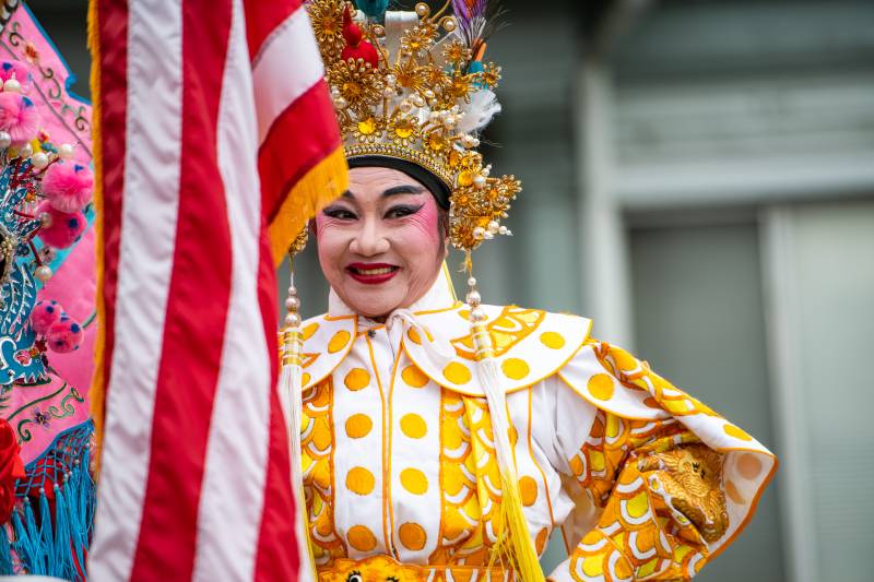 A person wearing an embroidered golden costume and headpiece stands next to an American flag outside.