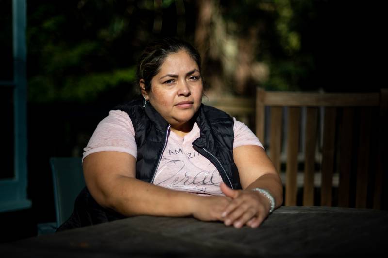 a portrait of a Latina woman in a pink shirt and vest with her hair pulled back, sitting with serious look on her face at an outdoor table