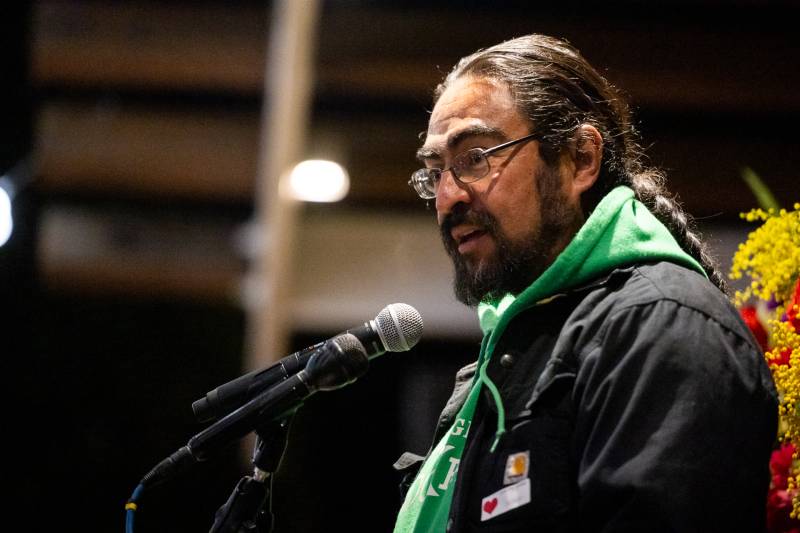 A Latino man with glasses and a beard wearing a green scarf, jacket, and with a pony tail, speaks into a microphone.