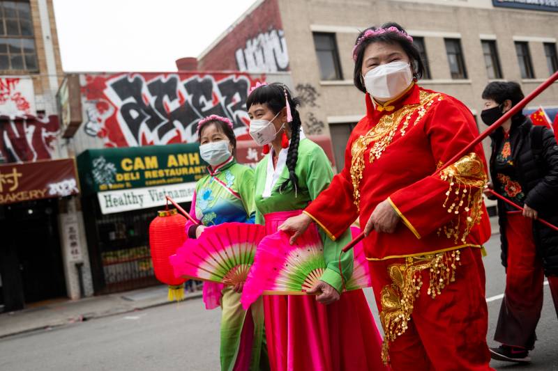 Three young women dressed in green and red costumes with face masks and holding fans walk on the street.