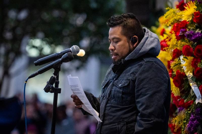 A Latino man speaks into a microphone as he reads from a sheet with flowers behind him.