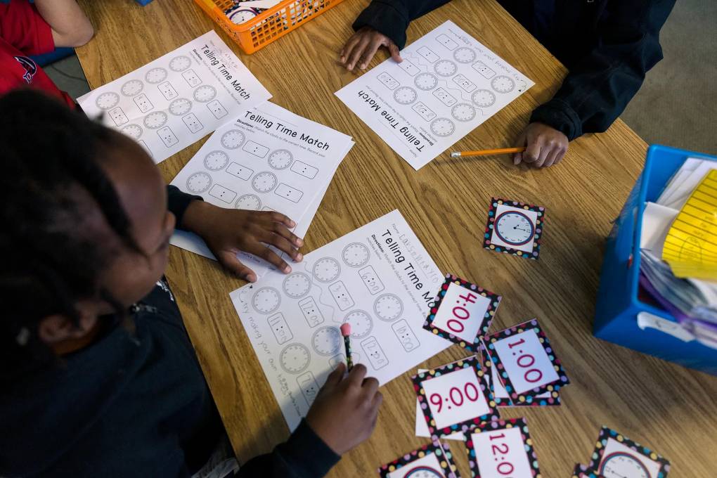 A downward-angled photo shows a school table with several students sitting around it, pencils in hand. The students are working on a worksheet about telling time.