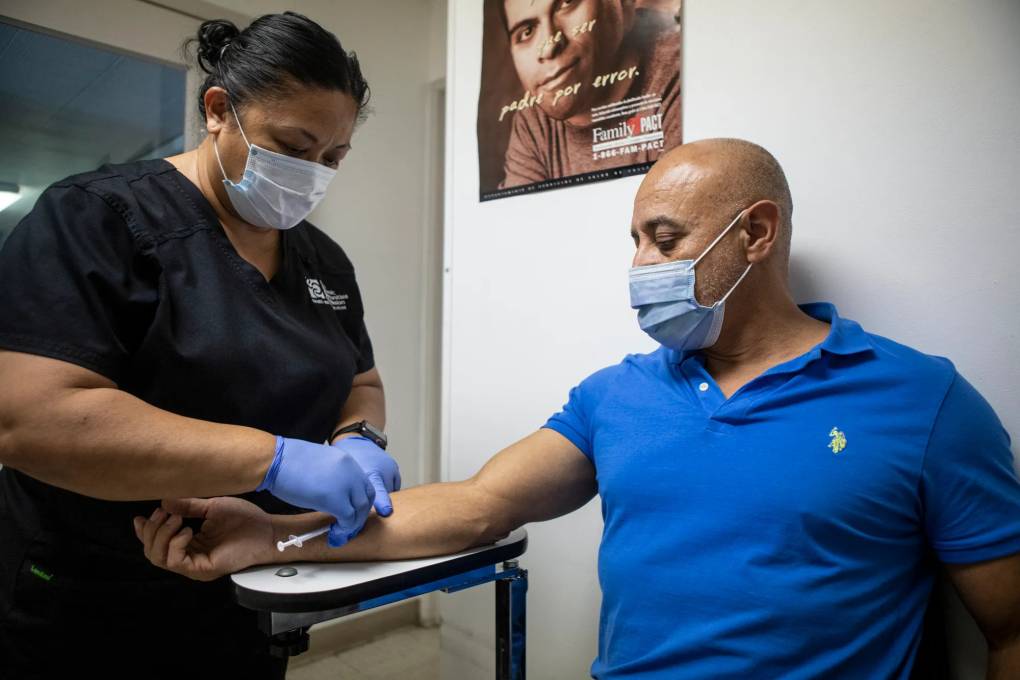 a man in a blue shirt wearing a mask receives a vaccine shot from a nurse with black hair in a dark blue shirt