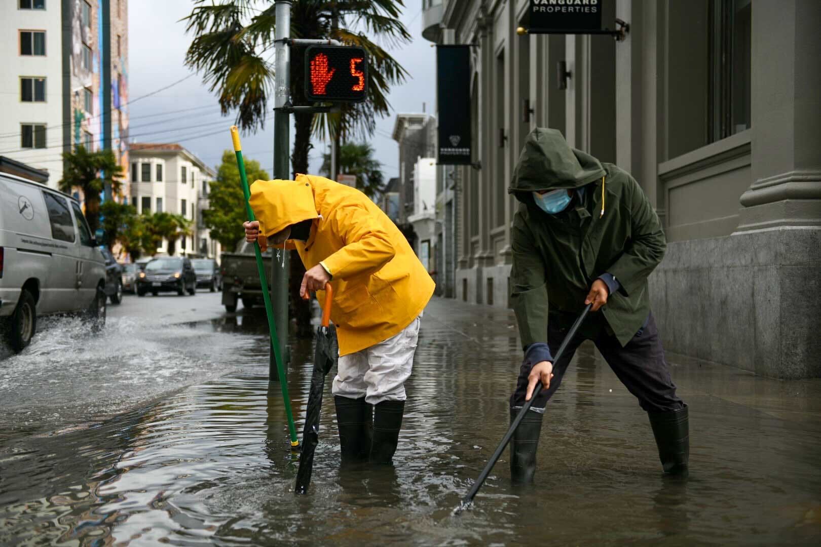 two people in raincoats stand in shin deep water as they try to clear a drain on a city street in San Francisco