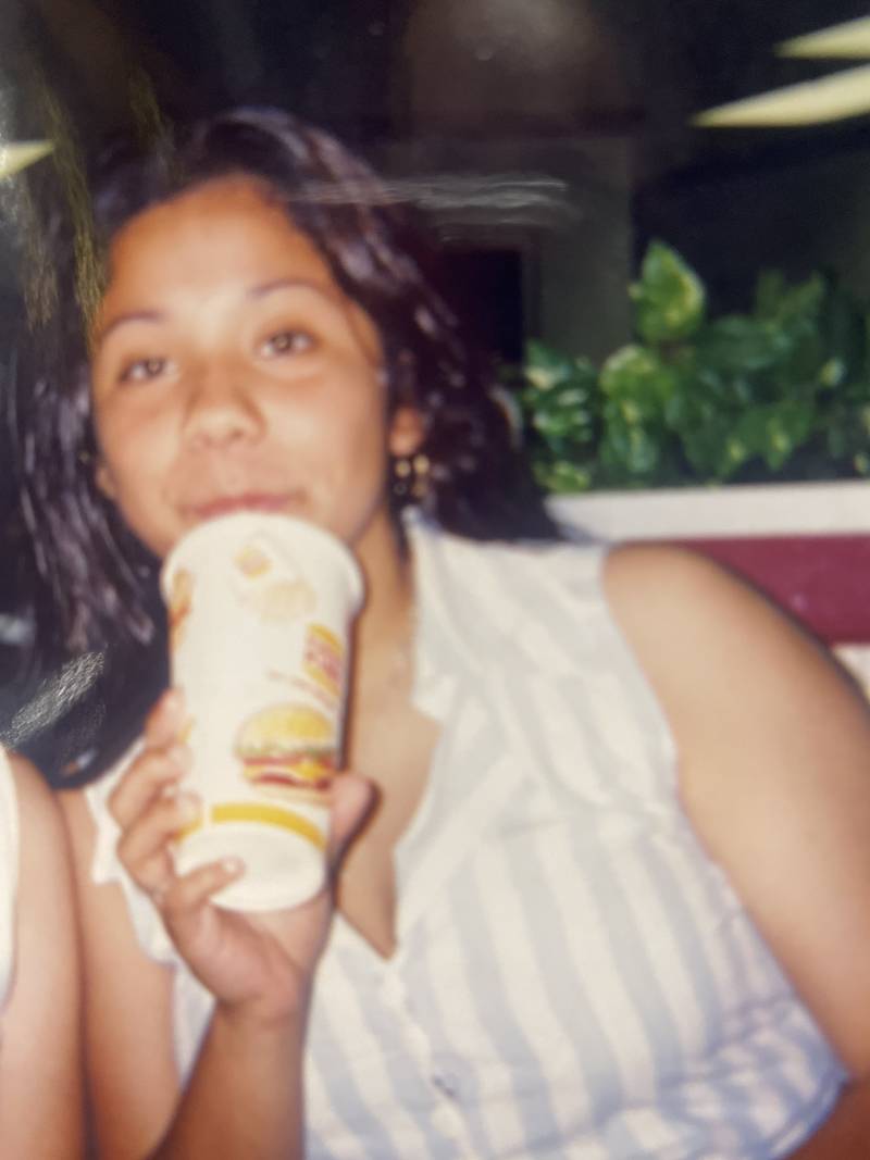 A Latina girl sips a soft drink with white dress on and long brown hair.
