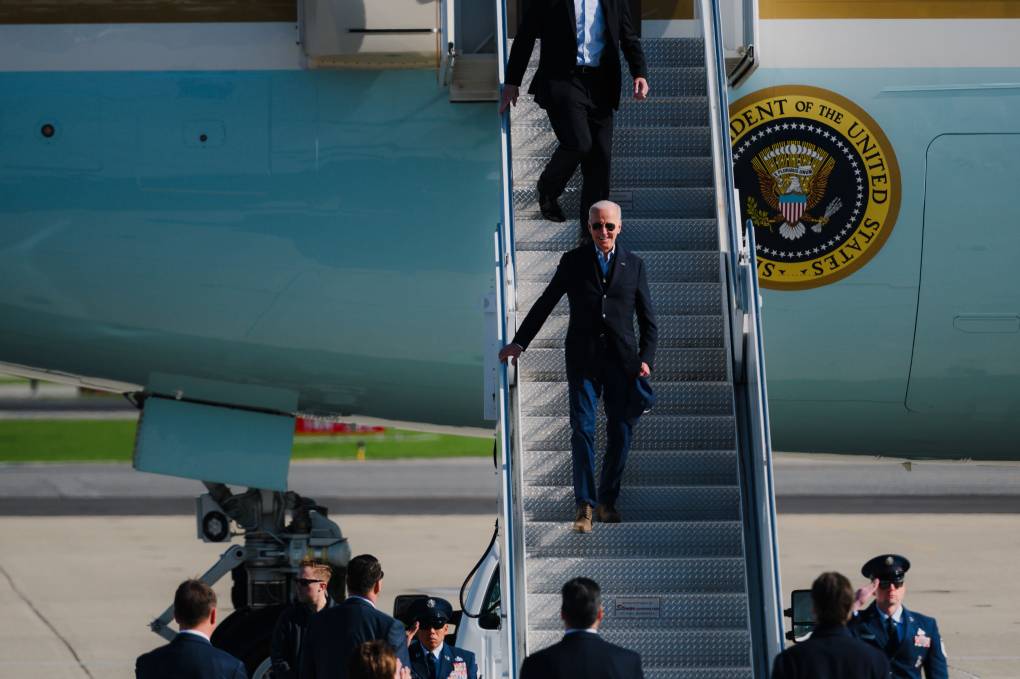 An elderly white man exits Air Force One with presidential seal on the airplane and journalists and camera people waiting below, a soldier salutes.