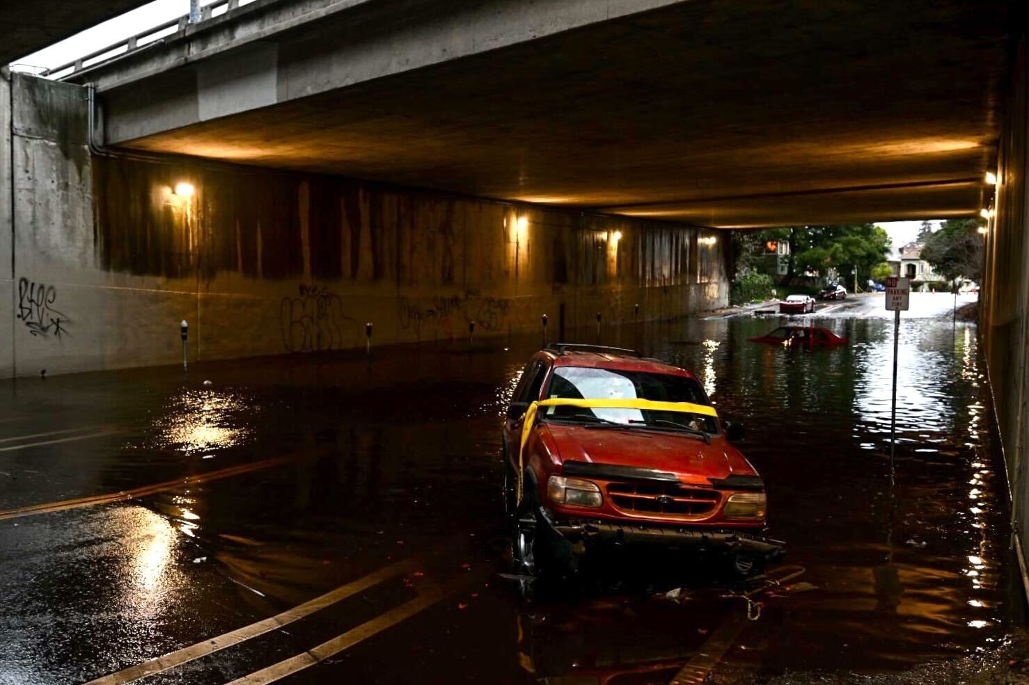 A red car lies abandoned under a flooded underpass with another car submerged behind it.