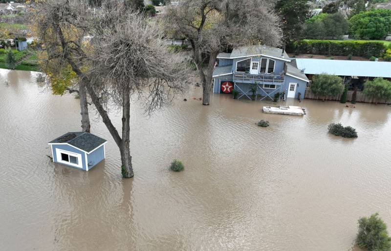 Two buildings are partially submerged in murky brown water. One small shack in the foreground, and a larger house in the background.