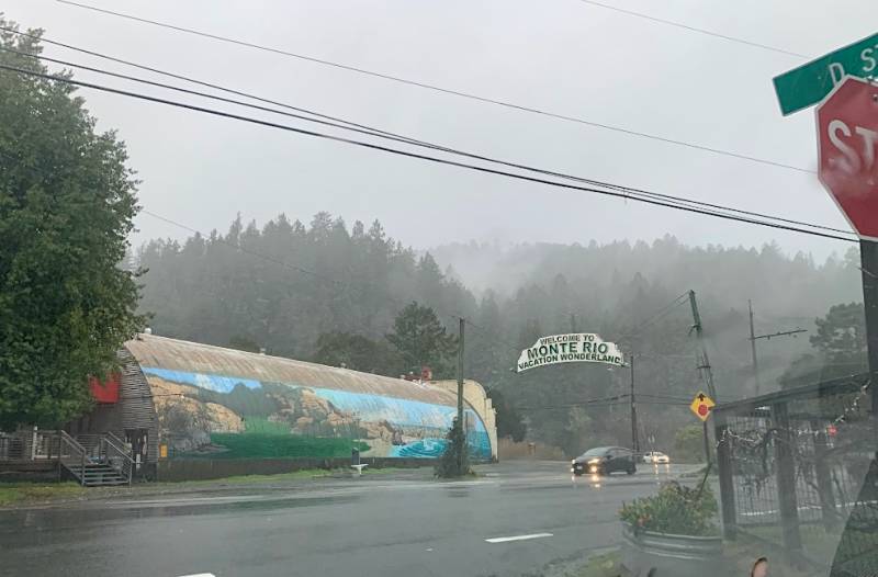 a stormy, rainy day with a grey sky in Monte Rio, a community on the Russian River. A sign says 'Welcome to Monte Rio, vacation wonderland' as a car drives through the storm with headlights on