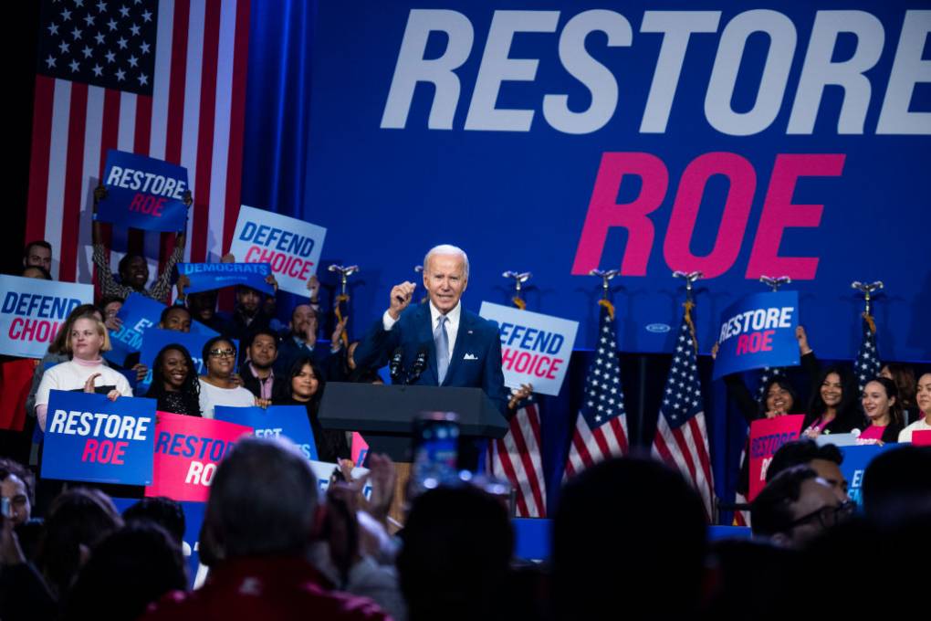 An octogenarian white man in a dark blue suit and light blue tie raises his fist as crowds around him cheer, with the words "Restore Roe" written in big letters behind him and crowds holding U.S. flags in the foreground.