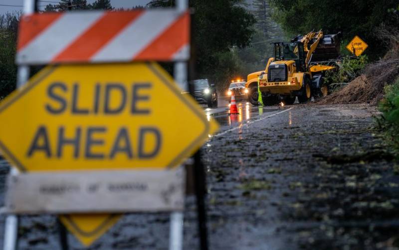 a sign that says 'slide ahead' is seen as bulldozer cleans up debris from a mudslide along a wet road as cars pass by with their headlights on