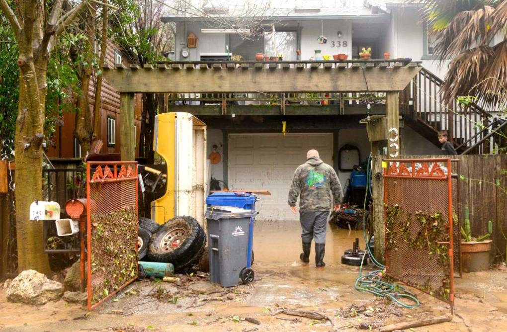 A man faces a home dressed in rain boots, grey pants and a camouflage hoodie surrounded by debris, tires, trash bins and muddy water.