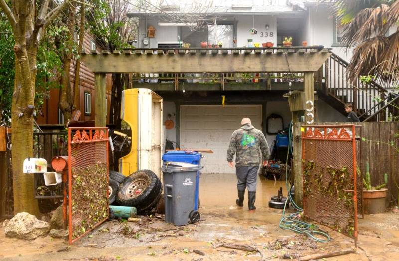 A man faces a home dressed in rain boots, grey pants and a camouflage hoodie surrounded by debris, tires, trash bins and muddy water.