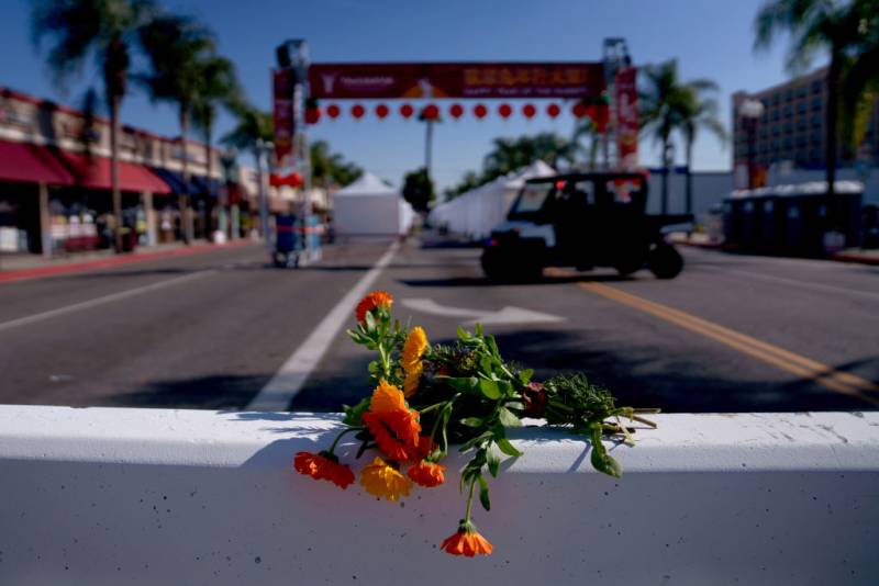 Orange flowers lie on a guard rail in the street with a vehicle and a red banner in the background.