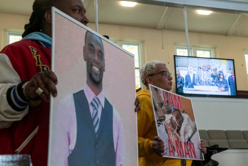 Two people, a Black man and a Black woman, hold signs with the image of Tyre Nichols with a TV in the background.