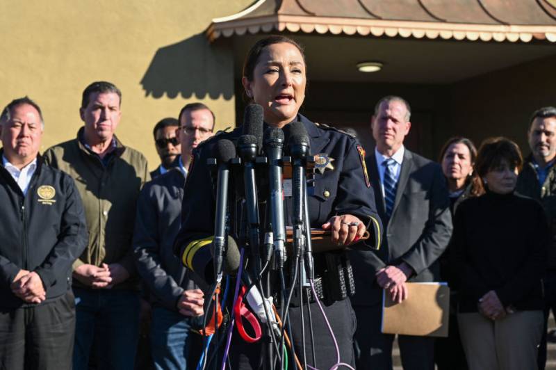 A female law enforcement officer speaks into microphones on an outdoor podium, with a group of men in suits standing behind her.
