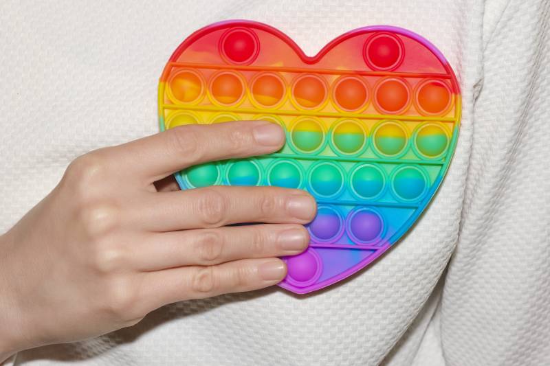 Woman holding rainbow-colored Pop Fidget Sensory Toy for Autism Special Needs Stress Relief in form of heart in her hand close up.