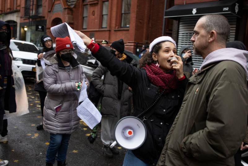 A woman of color with a megaphone gets close to the face of an angry stone-faced white man as she speaks into the megaphone