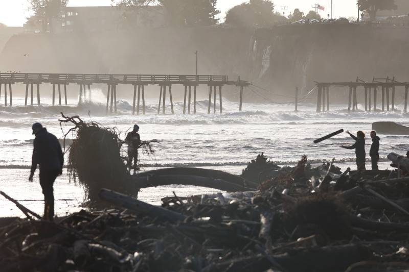 People gather near storm debris washed up on the beach, with a storm-damaged pier in the background.