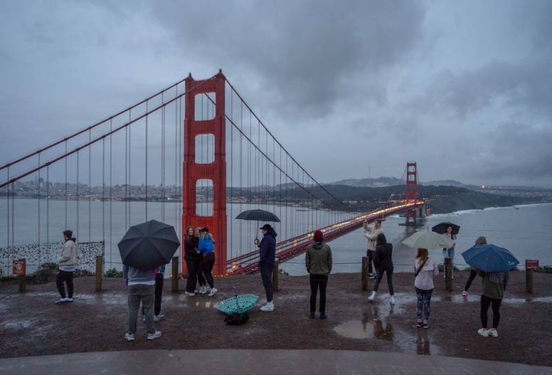 Several people dressed in raincoats and holding umbrellas stand behind the Golden Gate Bridge.