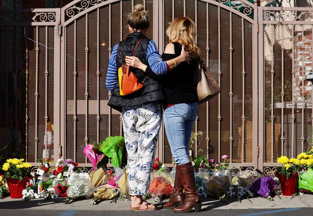 Two people stand side by side, arm in arm in front of a makeshift memorial. They face a gate with the sidewalk in front filled with flowers.