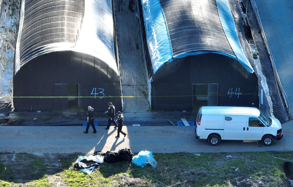 An aerial phot of police walking past several large warehouse buildings that are cordoned off with caution tape.