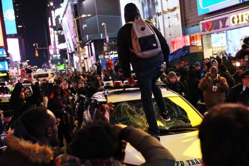 A man stomps on a police car windshield in a crowd of people on a busy city street.