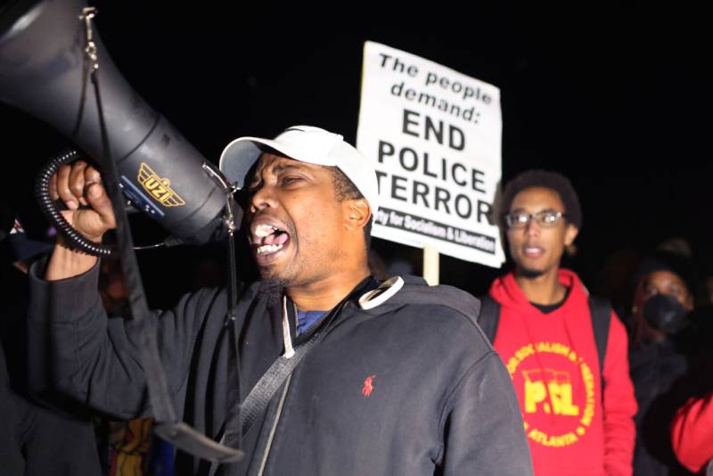 A young Black man speaks into a loudspeaker with a young Black woman standing behind him holding a sign that reads "Stop Police Terror."