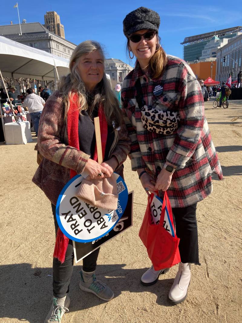 Two white woman, the shorter on the left with long, gray hair, and the taller and younger on the right with long brown hair, pose for a picture. The older woman has an uncertain look on her face and clasps and peach colored beanie and a protest sign against her body; the younger woman wears sunglasses and a black cap, wearing a plaid shirt and a leopard-spotted fanny pack slung across her chest, and she smiles broadly.