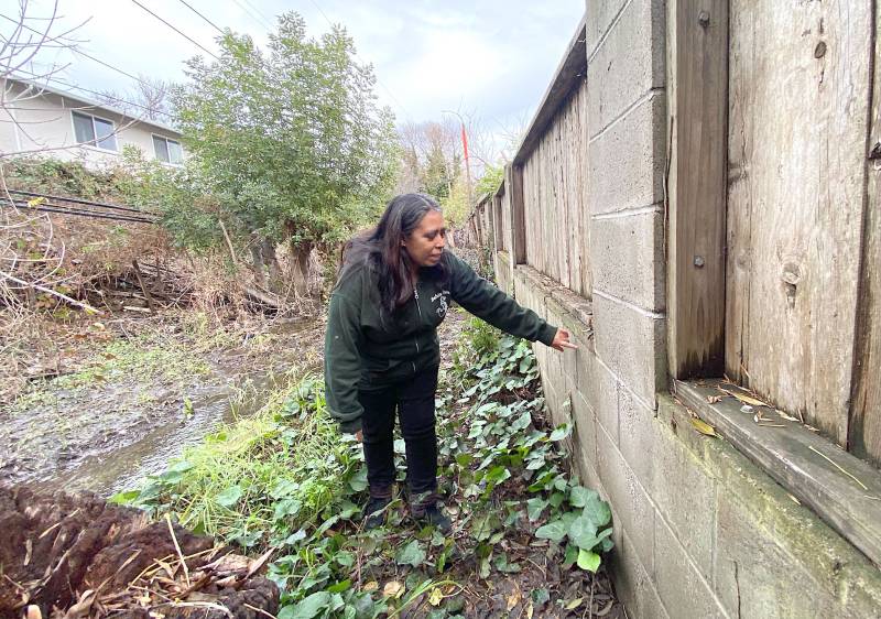 A woman stands by a creek, pointing to a cement wall.