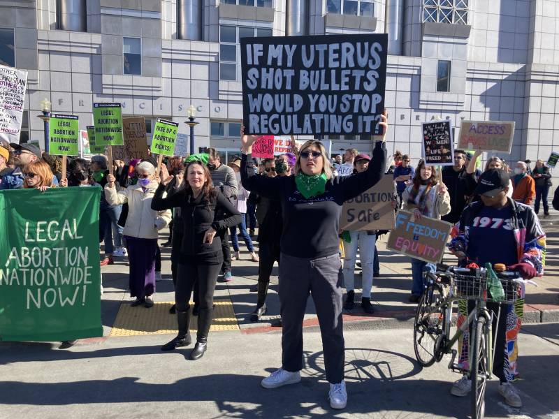 A woman stands in front of a group of abortion rights supporters holding a sign that reads "If my uterus shot bullets would you stop regulating it?"