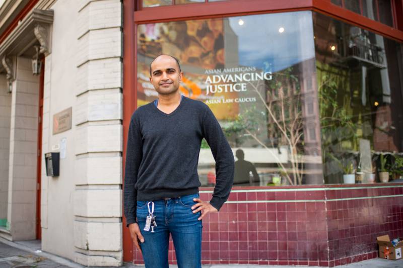 A south Asian man in a green sweater and jeans stands outside an office with "Advancing Justice" written on the window.