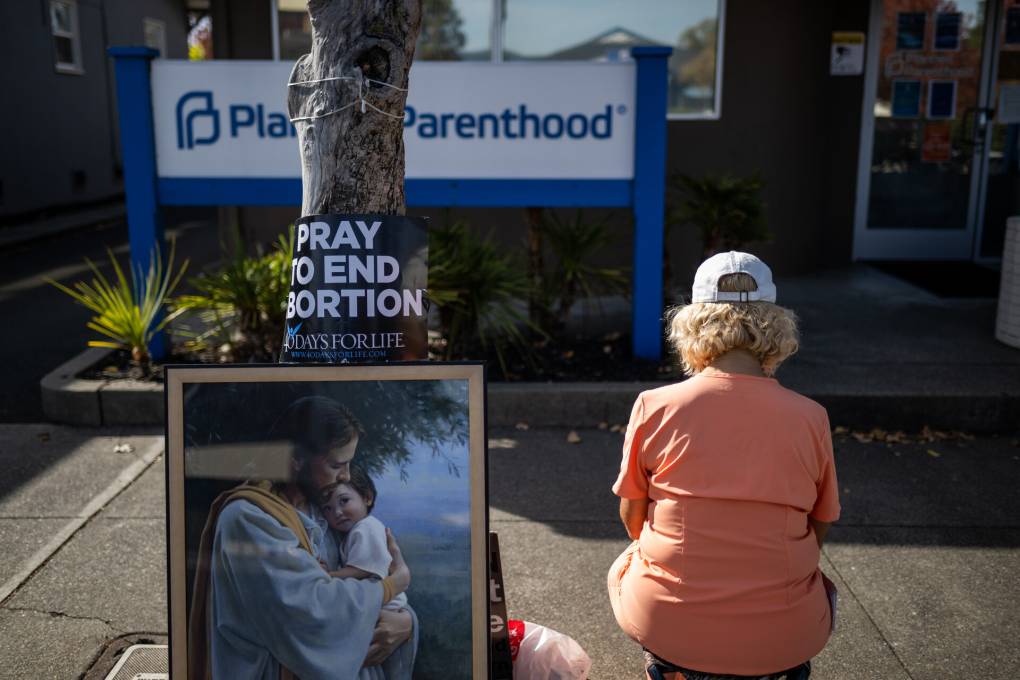 a woman with blonde hair is seen from behind sitting in orange scrubs outside a Planned Parenthood, next to a painting of Jesus and a sign that reads 'pray to end abortion'