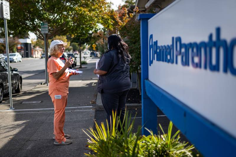 a older white woman in orange scrubs talks to a Black woman in dark clothing outside a Planned Parenthood health clinic