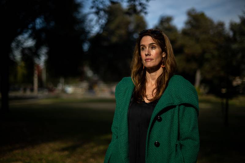 a woman with long brown hair wearing a green overcoat poses in a park