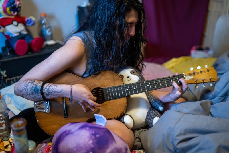 A Caucasian with long dark hair, wearing a sleeveless top and a tattoo on his right forearm, sits on a bed surrounded by stuffed animals and colorful cushions, playing a guitar.