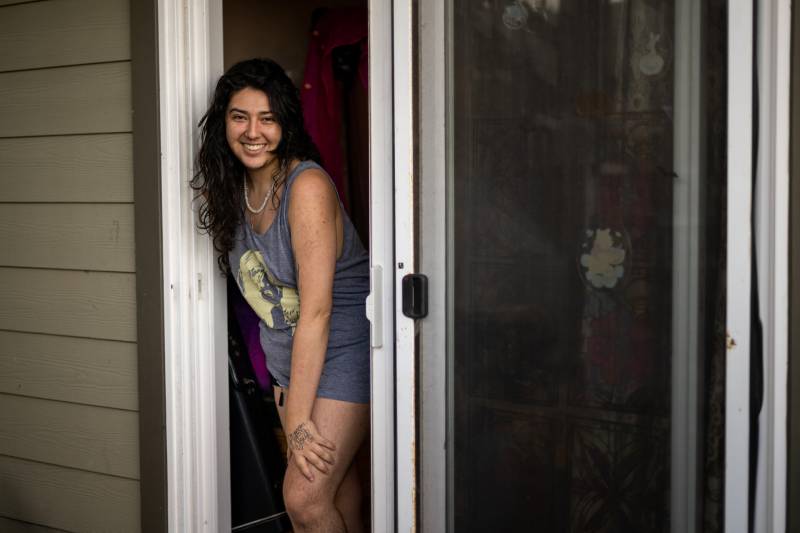 A transgender person with long dark hair and a shirt-dress smiles from their door at the camera.