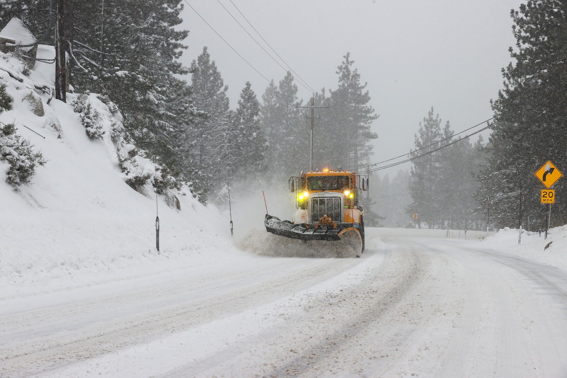 A mostly white photo, of a two-lane road covered in white and gray snow, penned in by snowy embankments on both sides with conifers, and an orange truck with bright yellow headlights clearing the road on the left side, facing the camera.