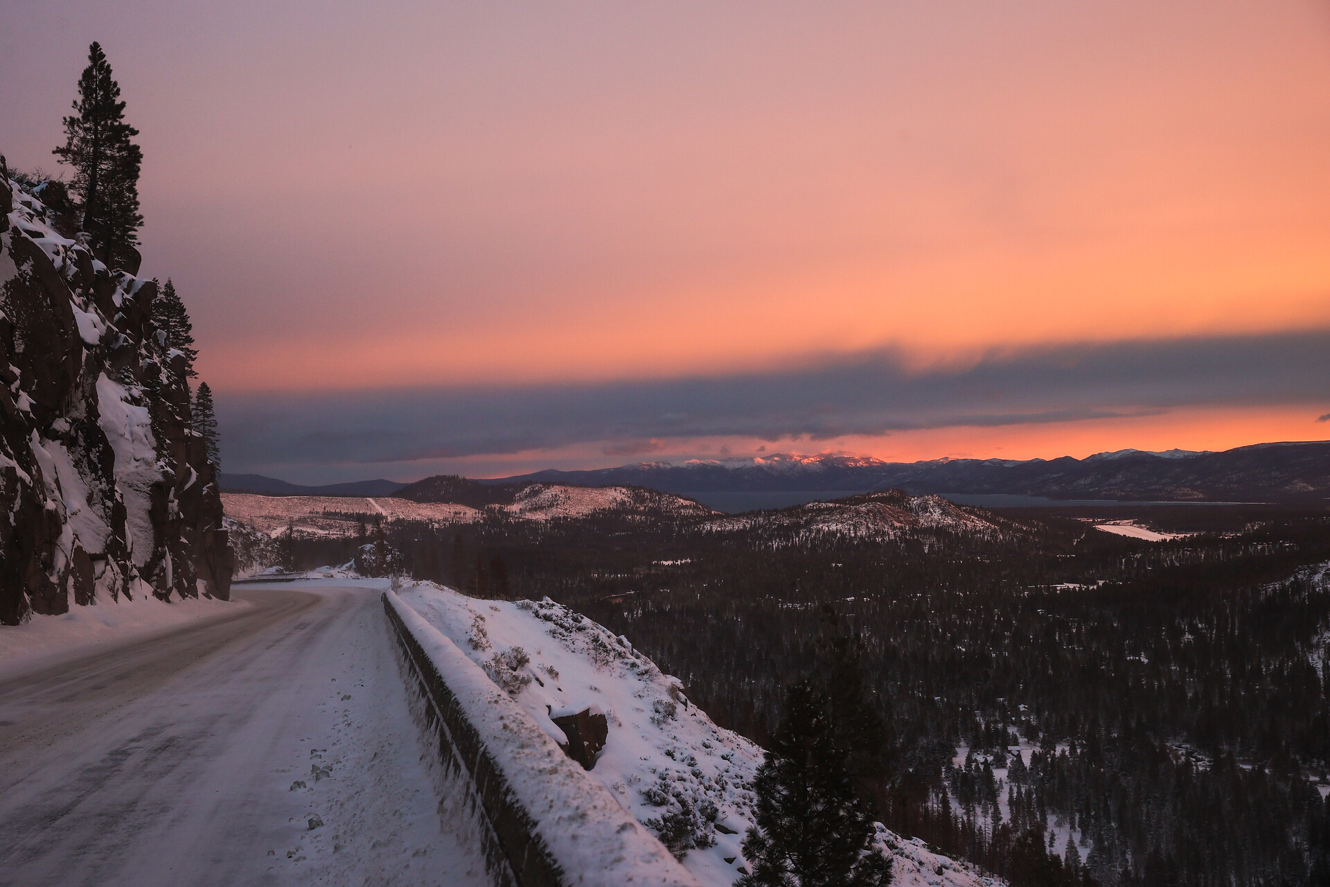 A peachy pink sky, with a low, blue line of clouds, taken from the side of a snowy road overlooking a valley full of conifers and snow.