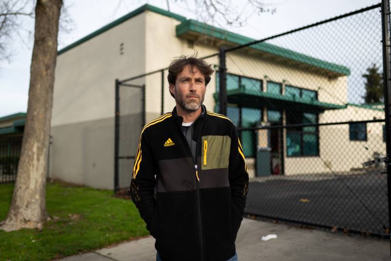 A man wearing a black track suit top with yellow stripes on the sides stands outside a building.