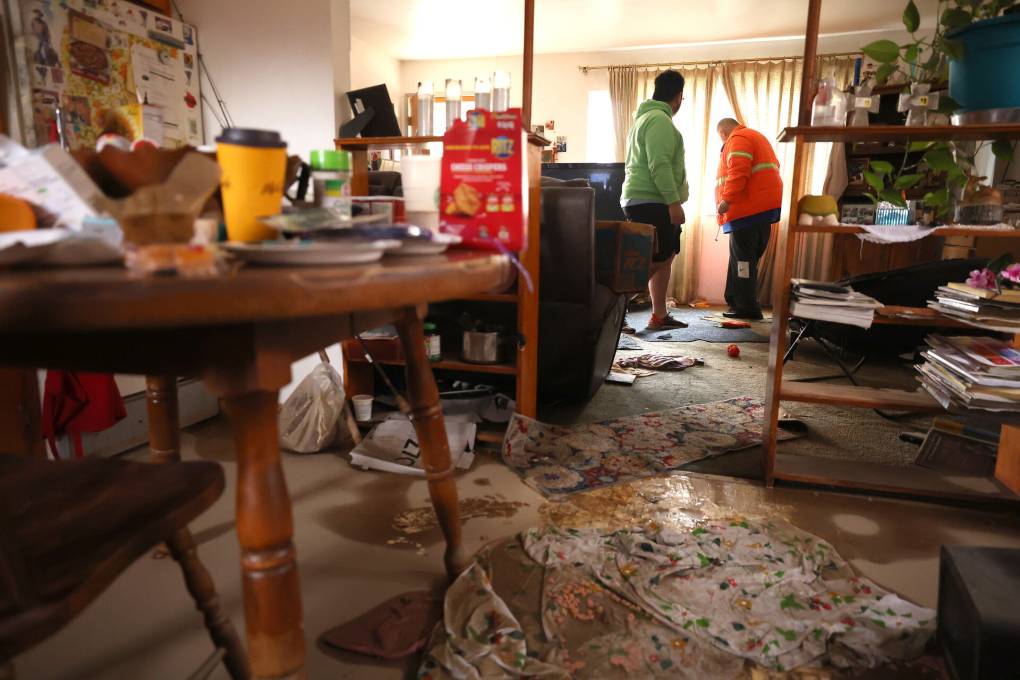A photograph showing the interior of a home, probably a living room, that has been damaged by flooding. Two people are in the back of the photo, examining the damage.
