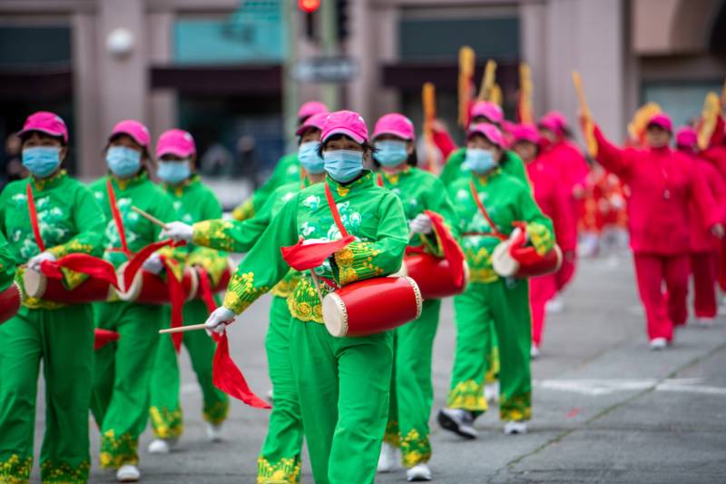 People dressed in bright green costumes and pink hats with face masks walk in formation while holding red drums and drum sticks.