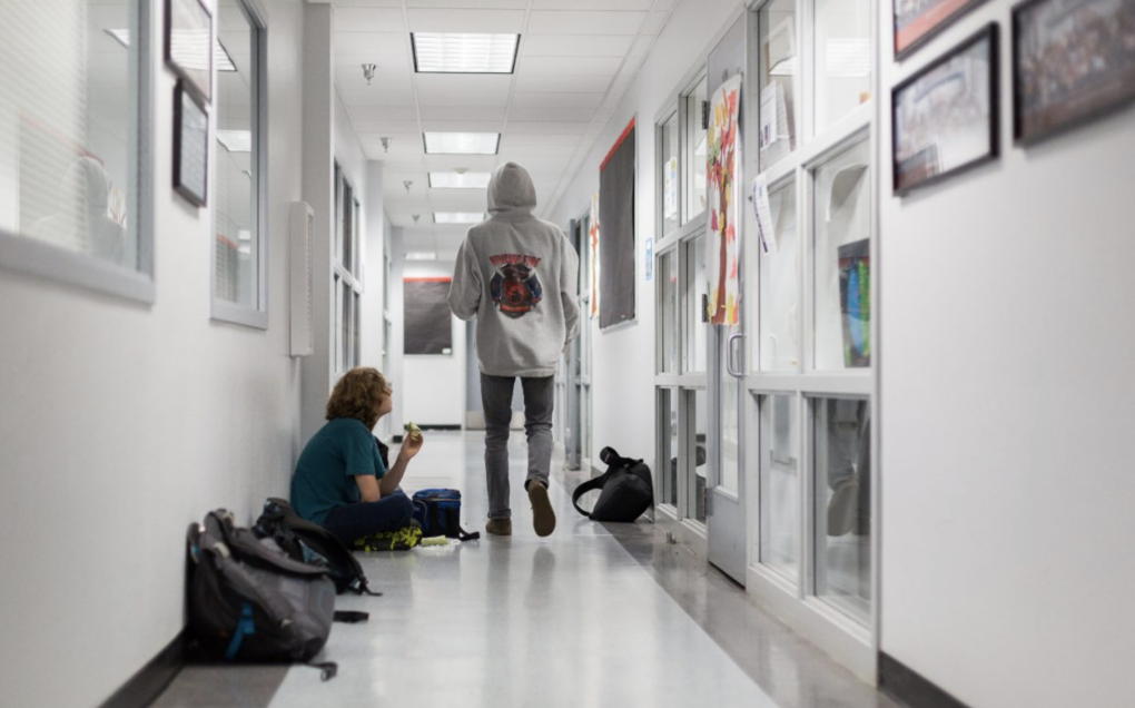 A young person walking down a hallway with a hood over their head while other young people sit down next to backpacks.